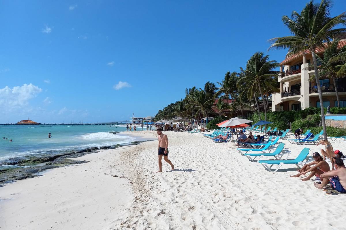 Vit sandstrand med solstolar, parasoller och palmer framför låga hotell vid turkost vatten i Playa del Carmen