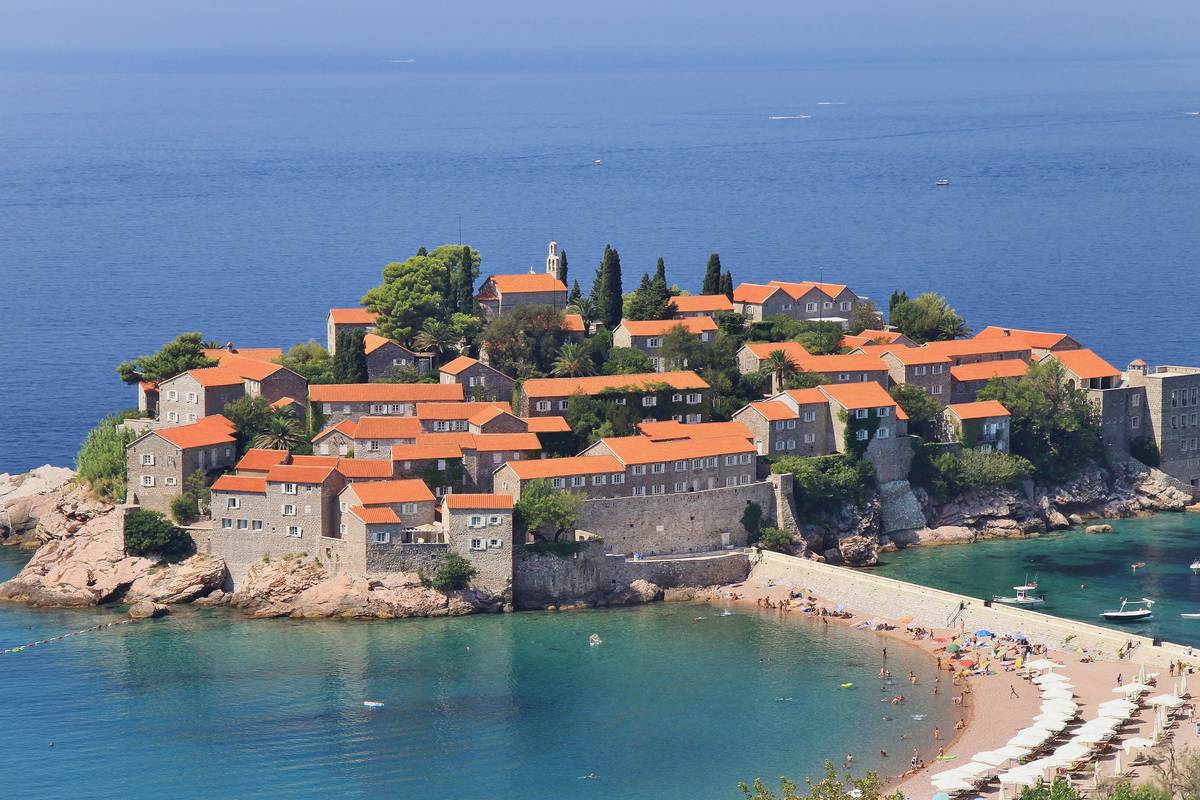 Ön Sveti Stefan med stenhus och orange tak på en klippa i det turkosa havet, förbunden med land av en ljus sandremsa med strand