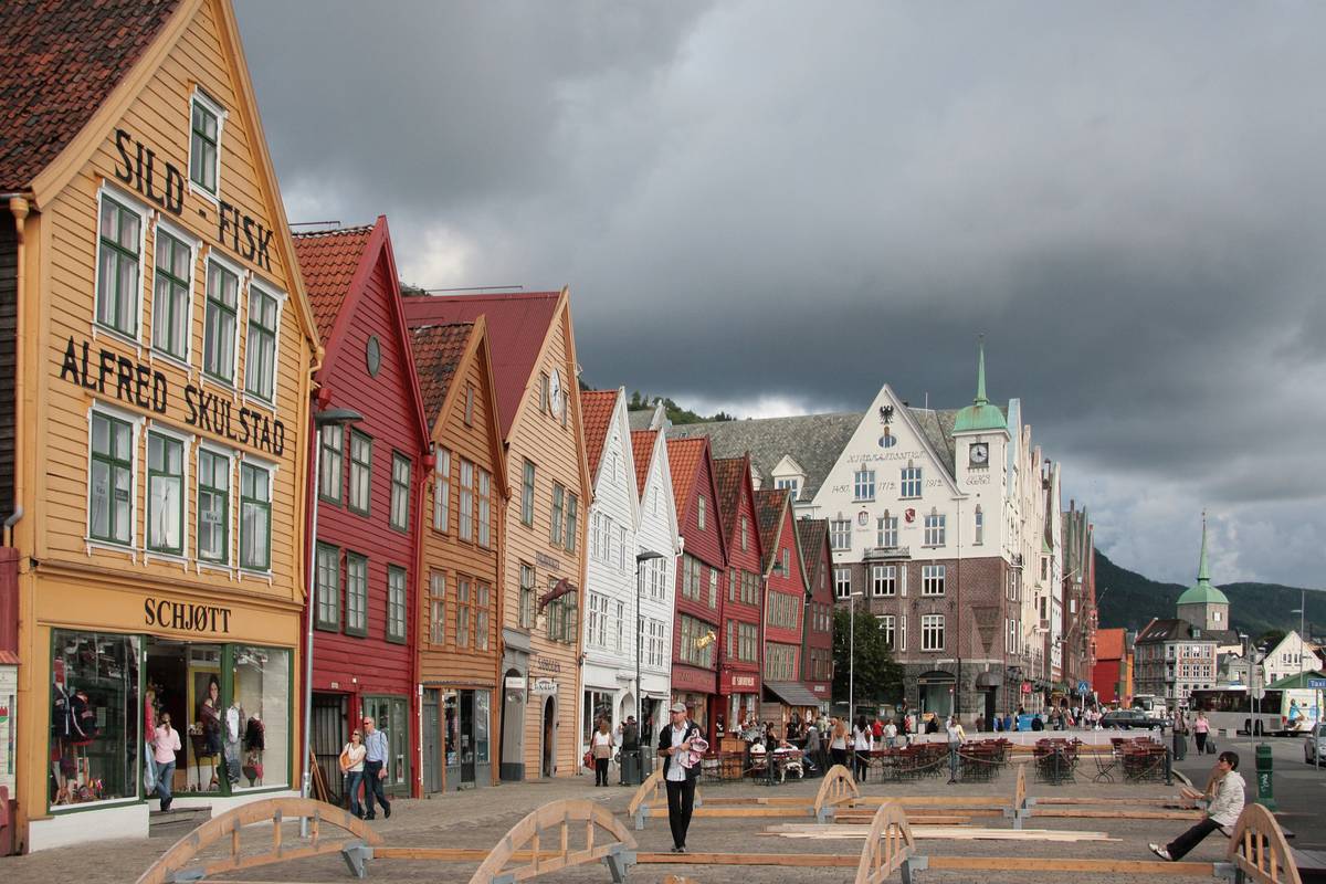 Raden av färgglada trähus med spetsiga gavlar på Bryggen i Bergen, med människor som promenerar på kajen under en mulen himmel