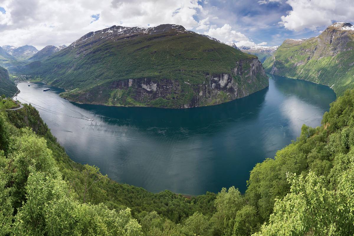 Utsikt från Ørnesvingen över den djupblå, slingrande Geirangerfjorden mellan branta, gröna berg med lövskog i förgrunden och snöfläckar på topparna