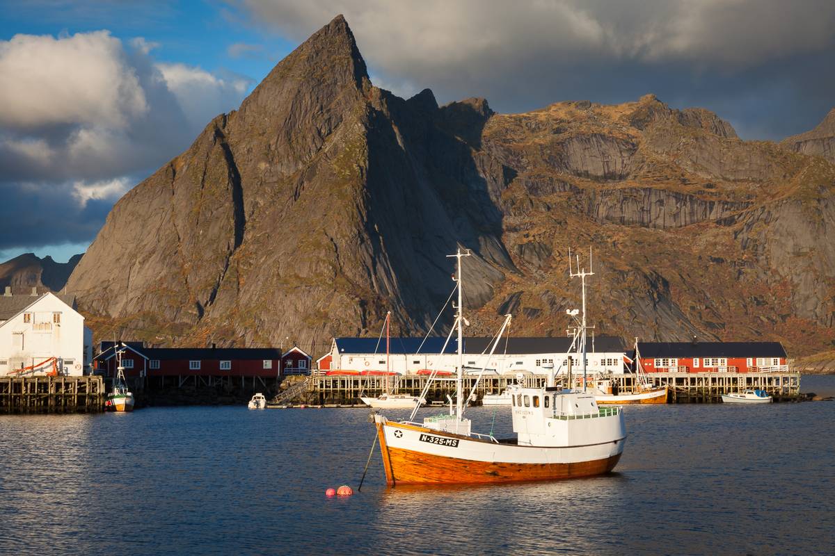 En vit och orange fiskebåt i hamnen i Hamnøy nära Reine, med röda rorbuer på kajen och ett spetsigt, brant berg i kvällssol bakom