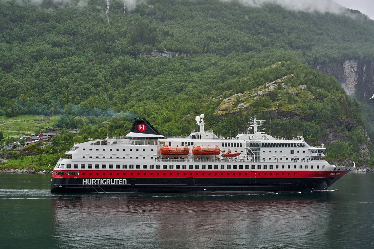 Hurtigrutens vit- och rödmålade fartyg Polarlys glider på Geirangerfjorden framför en grön, skogklädd bergssluttning