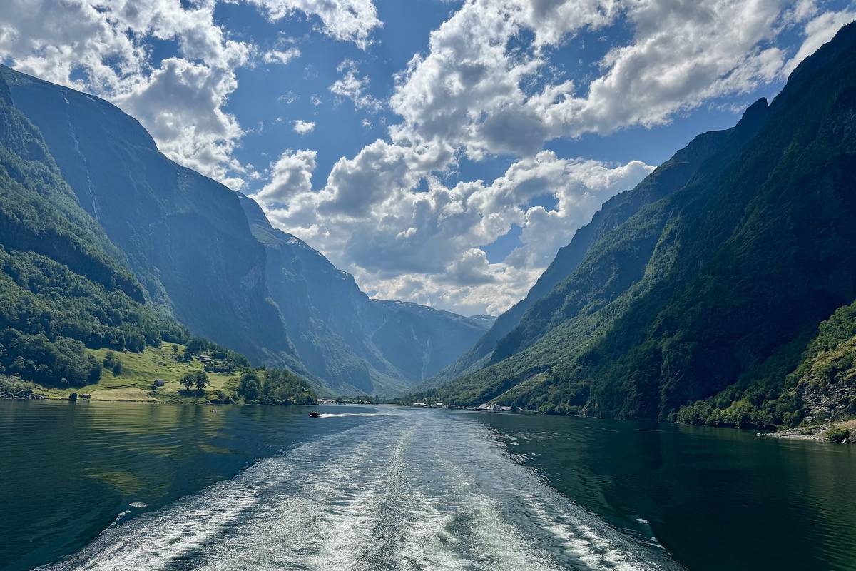 Kölvattnet från en båt på den smala Nærøyfjorden, med branta, gröna berg på båda sidor och vita moln på en blå himmel