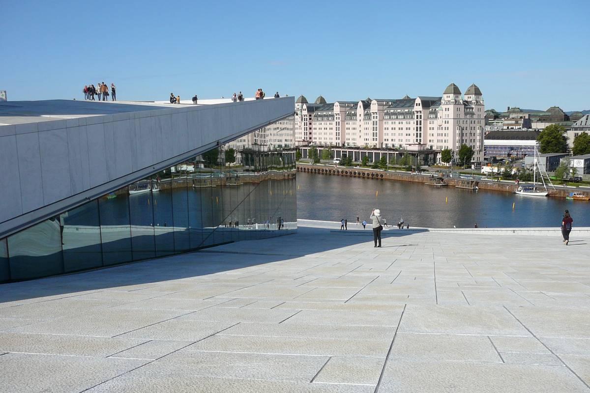 Människor promenerar på det sluttande vita marmortaket på Operan i Oslo, med vattnet och en stor vit byggnad i bakgrunden under en klarblå himmel