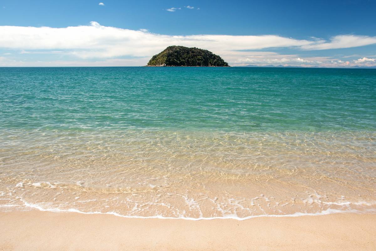 Gyllene sandstrand och klart turkost vatten i Abel Tasman nationalpark, med den skogklädda ön Tonga Island ute i havet