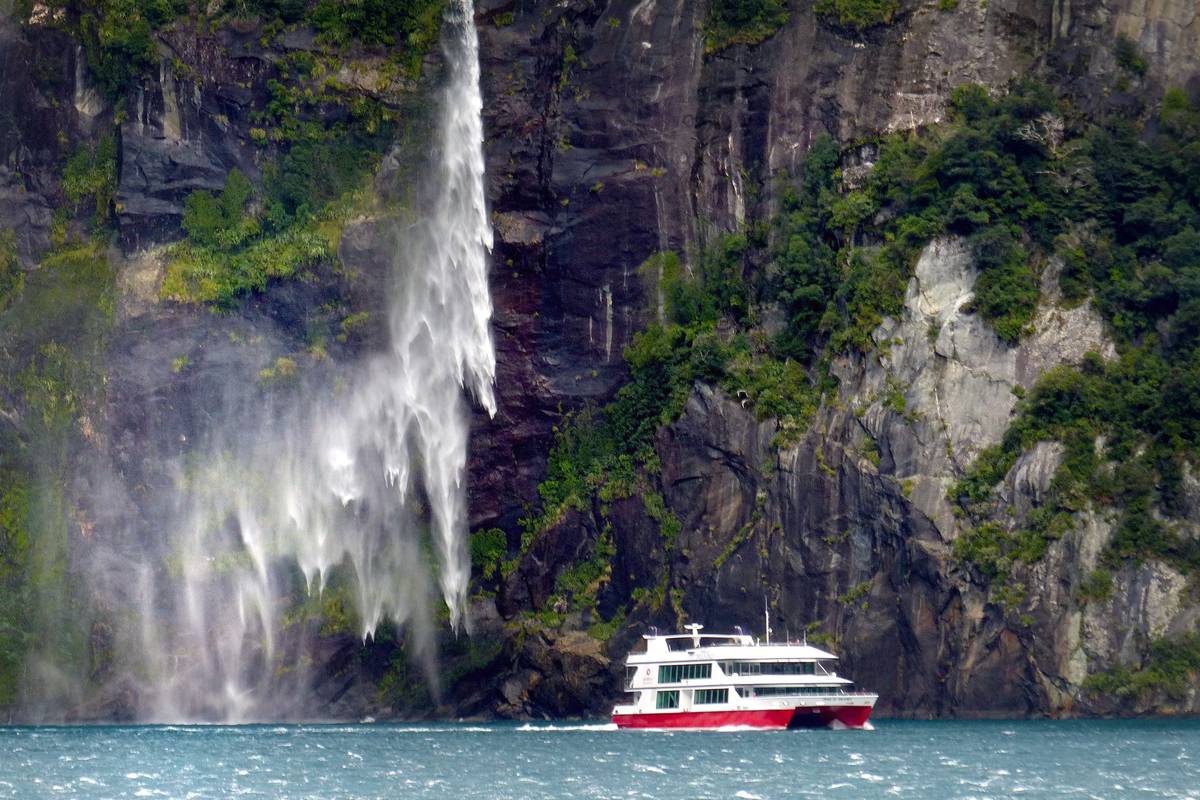 En vit och röd kryssningsbåt passerar under ett högt vattenfall som störtar ner för en mörk bergvägg i Milford Sound