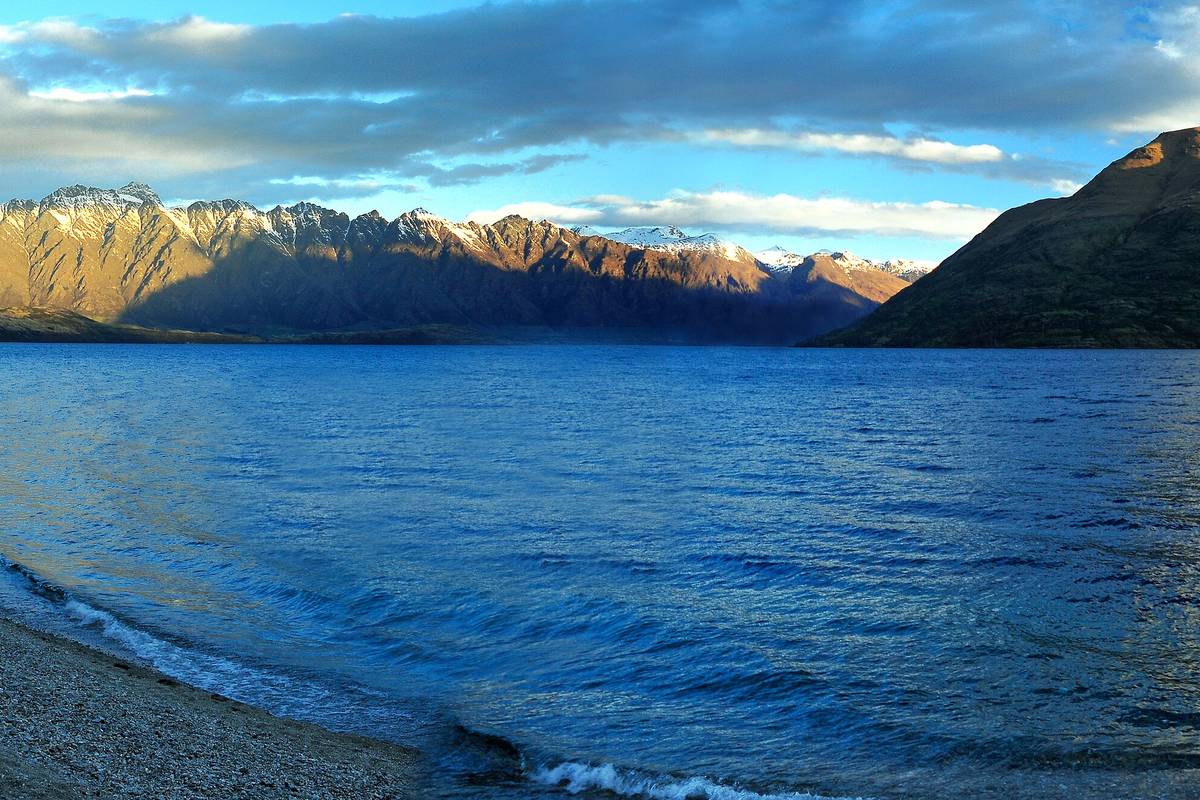 Lake Wakatipu i kvällsljus med bergskedjan The Remarkables i bakgrunden och en grusstrand i förgrunden