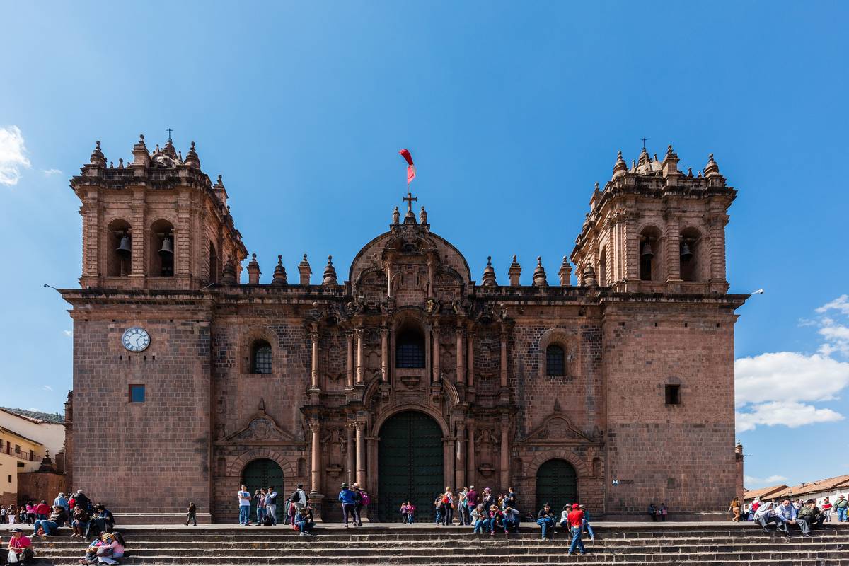 Katedralen i Cusco med två klocktorn i rödbrun sten vid Plaza de Armas under en klarblå himmel, med människor på trapporna
