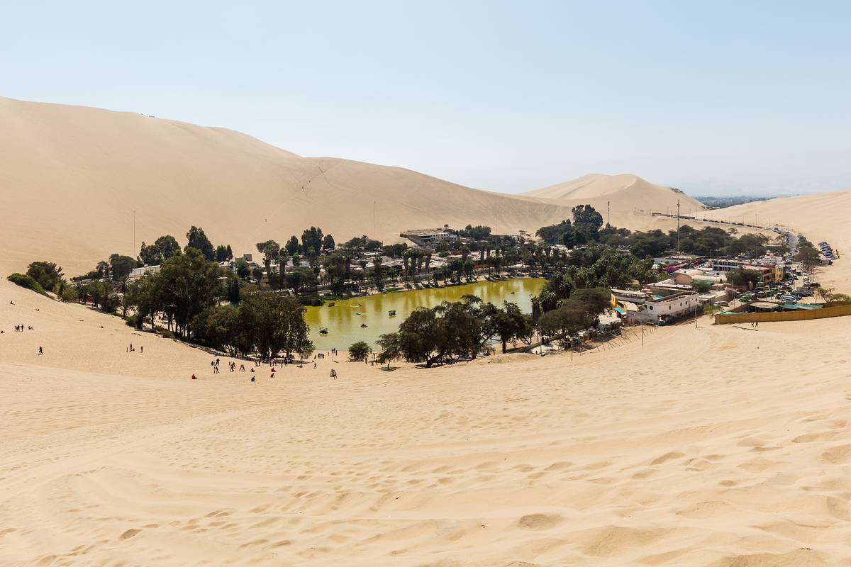 Oasen Huacachina med en grön lagun och palmer omgiven av höga, ljusa sanddyner under en dimmig himmel