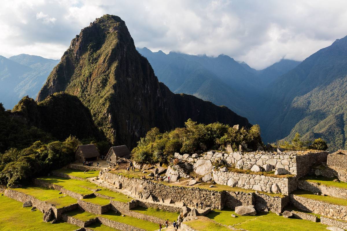 Gröna gräsklädda terrasser och stenhus i Machu Picchu framför det branta, skogsklädda berget Huayna Picchu och moln över bergen