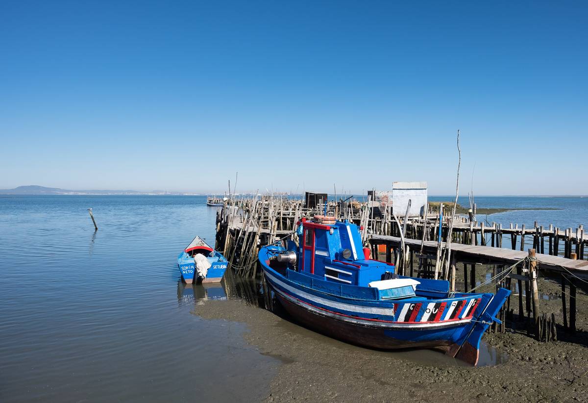 En blå fiskebåt vid fiskebryggan på träpålar i Carrasqueira nära Comporta, med lugnt vatten och klarblå himmel