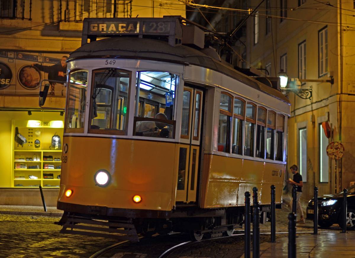 En gul spårvagn på linje 28 mot Graça på en kullerstensgata i Lissabon en kväll, med upplysta fönster och gamla husfasader