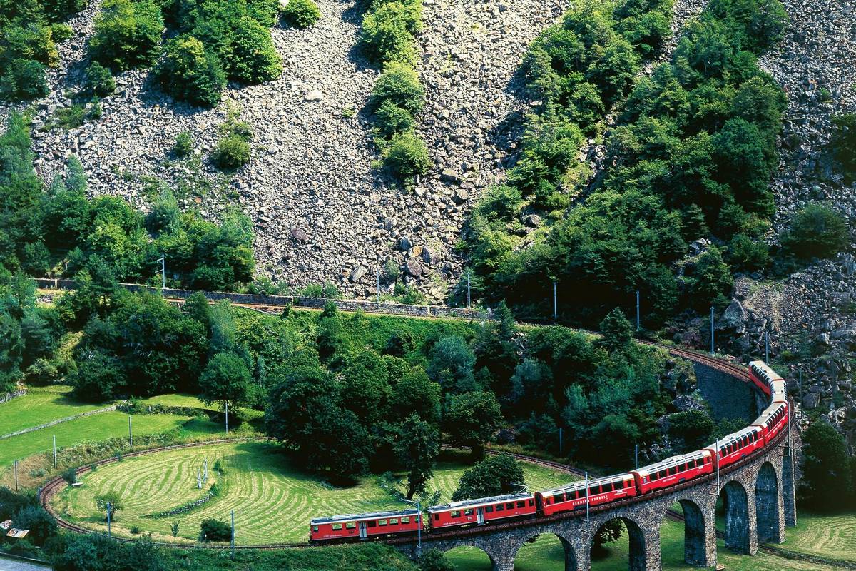 Ett rött Bernina Express-tåg kör i en spiral över den cirkelformade stenviadukten i Brusio, omgiven av gröna ängar, lövträd och en stenig sluttning