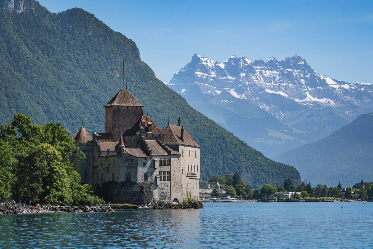 Den medeltida borgen Château de Chillon med torn och tegeltak på en klippa i Genèvesjön, med gröna berg och de snöklädda Dents du Midi i bakgrunden