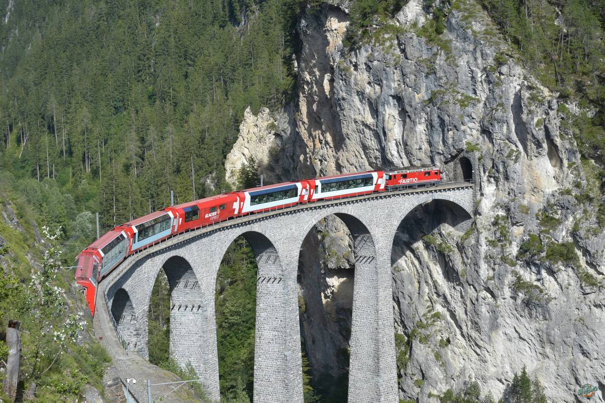 Ett rött Glacier Express-tåg med panoramavagnar kör över den höga, bågformade stenbron Landwasserviadukten och in i en tunnel i en lodrät klippvägg omgiven av skog