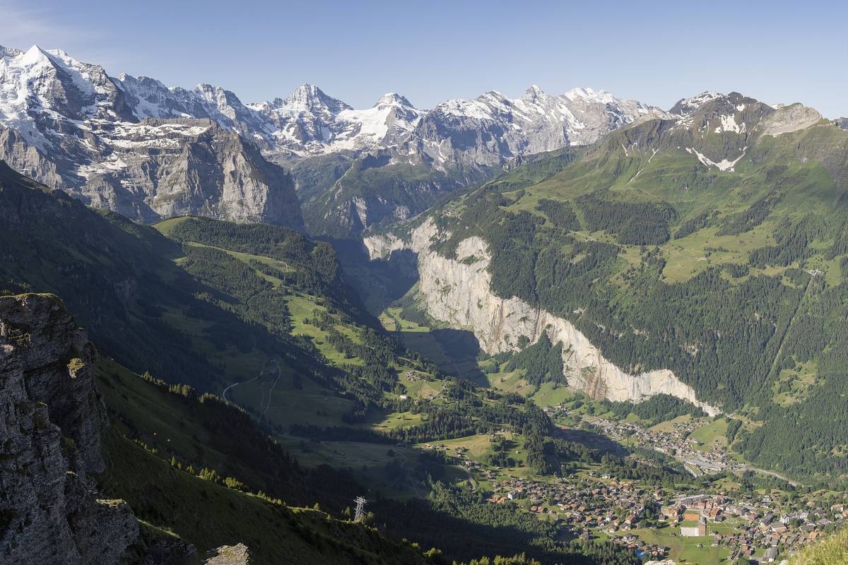 Utsikt från Wengen över den djupa, gröna Lauterbrunnendalen med branta klippväggar, vattenfall och snöklädda alptoppar under en klarblå himmel
