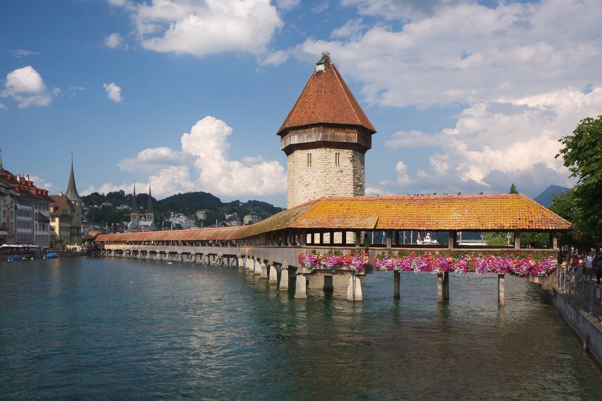 Den överbyggda träbron Kapellbrücke med röda blommor längs räcket och det åttkantiga stentornet Wasserturm i floden Reuss i Luzern, under en blå himmel med vita moln