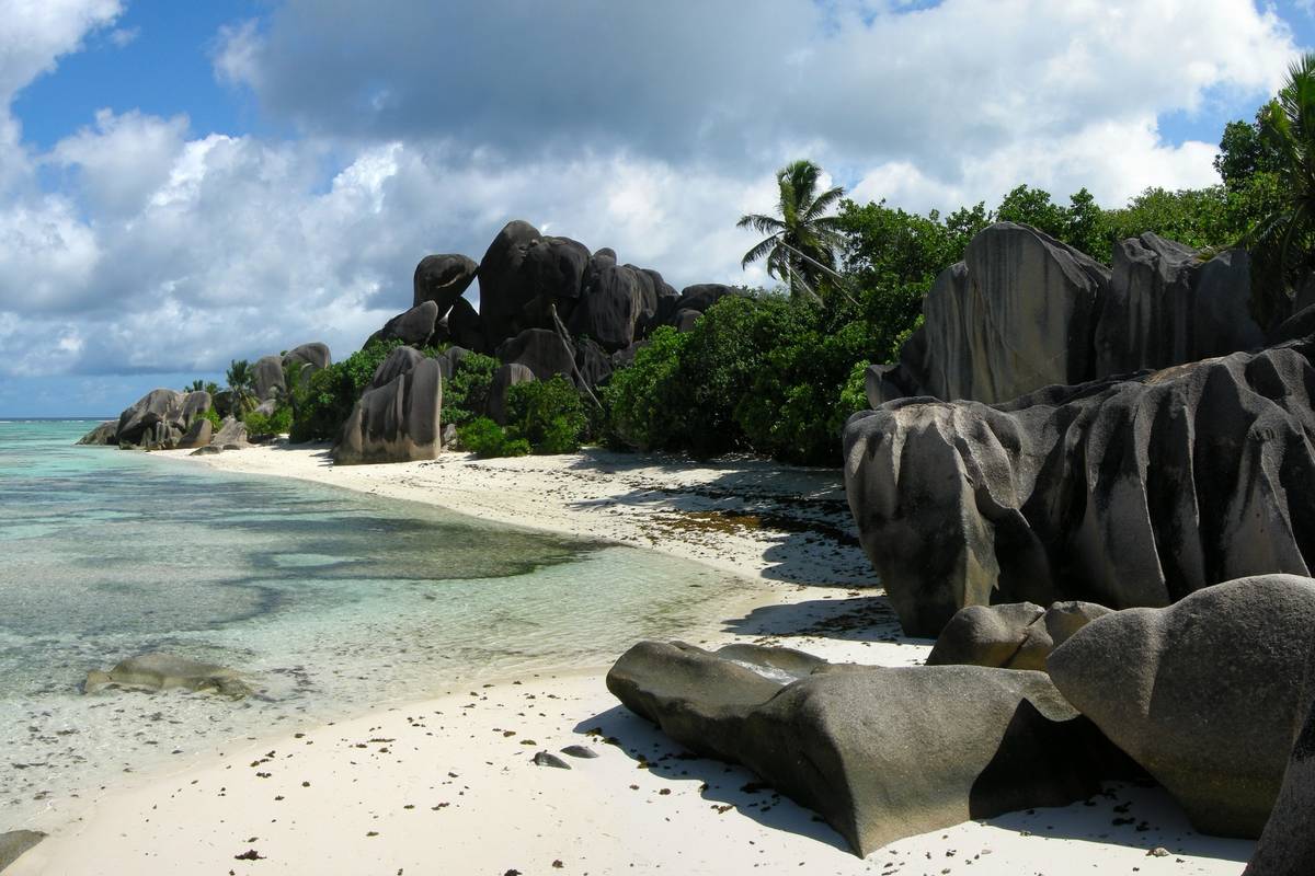 Mörkgrå, vattenslipade granitklippor med räfflor vid en vit sandstrand med grunt klart vatten, palmer och grönska vid Anse Source d'Argent