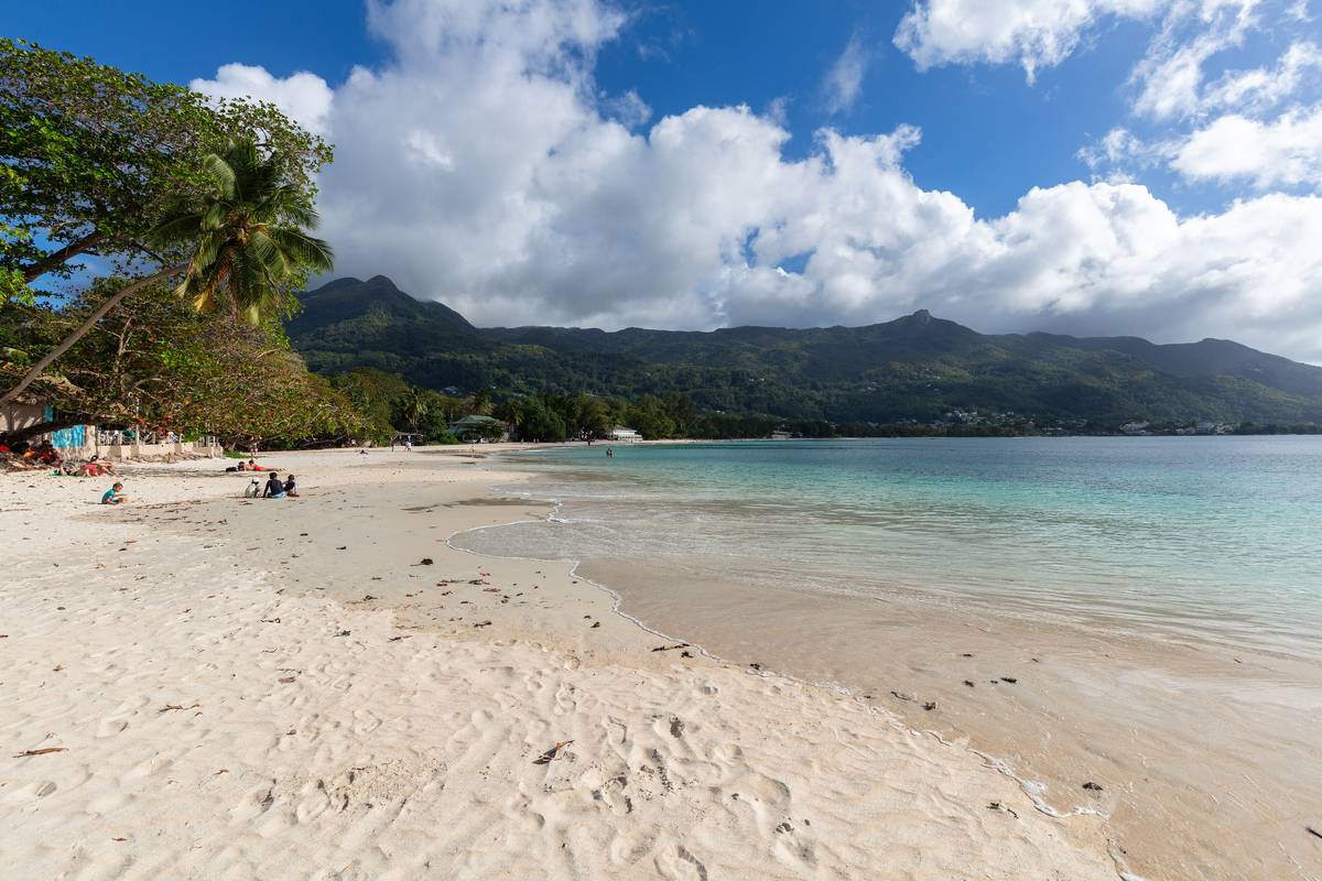 Den breda sandstranden Beau Vallon på Mahé med grunt, ljust turkost vatten, några badgäster och höga gröna berg under vita moln i bakgrunden