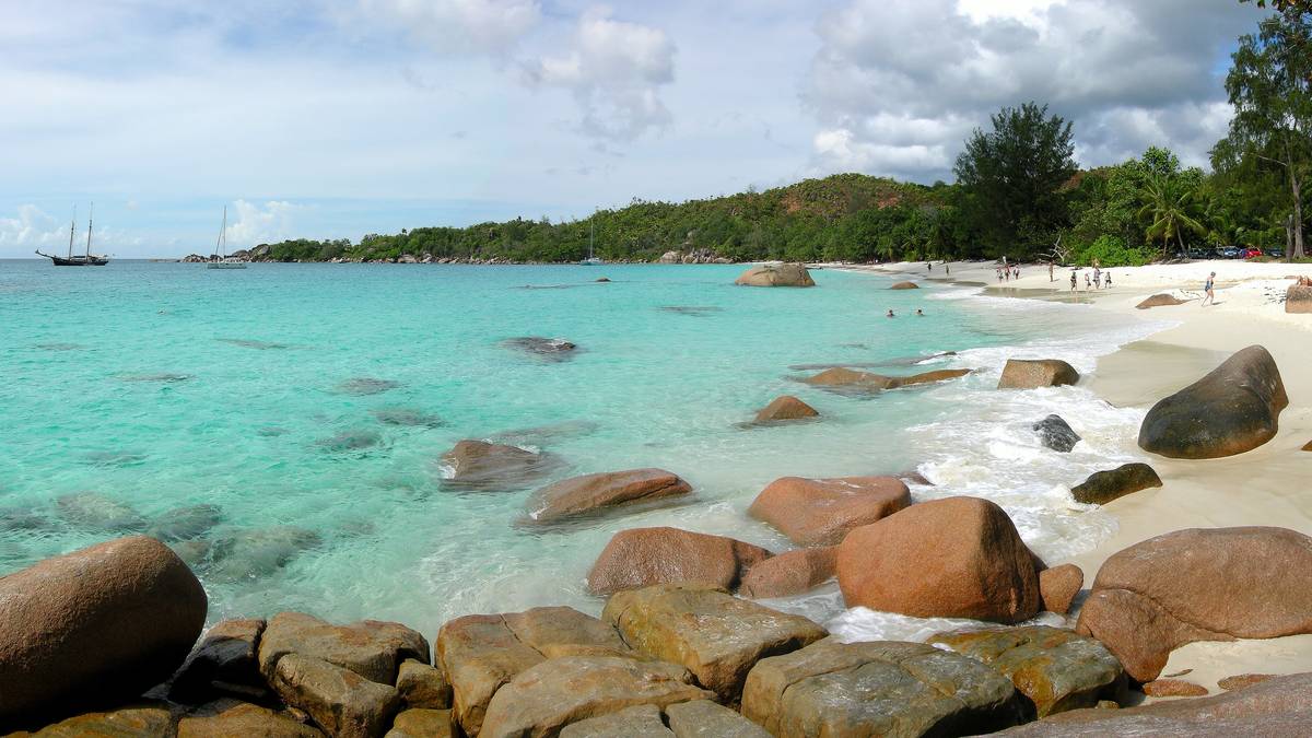 Rundade rödbruna granitblock i kristallklart turkost vatten vid Anse Lazio, med en lång vit sandstrand, gröna träd och segelbåtar i bakgrunden