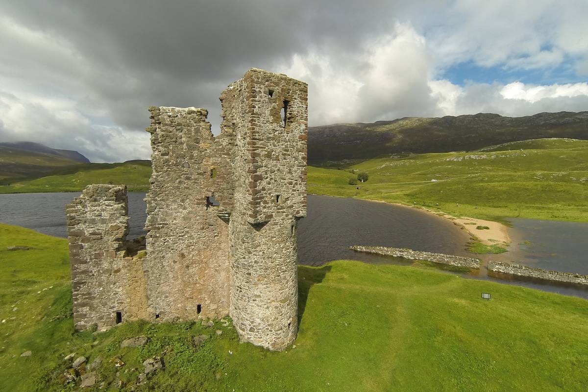 Den raserade stenborgen Ardvreck Castle på en grön udde i sjön Loch Assynt, med gröna kullar och mörka moln i bakgrunden