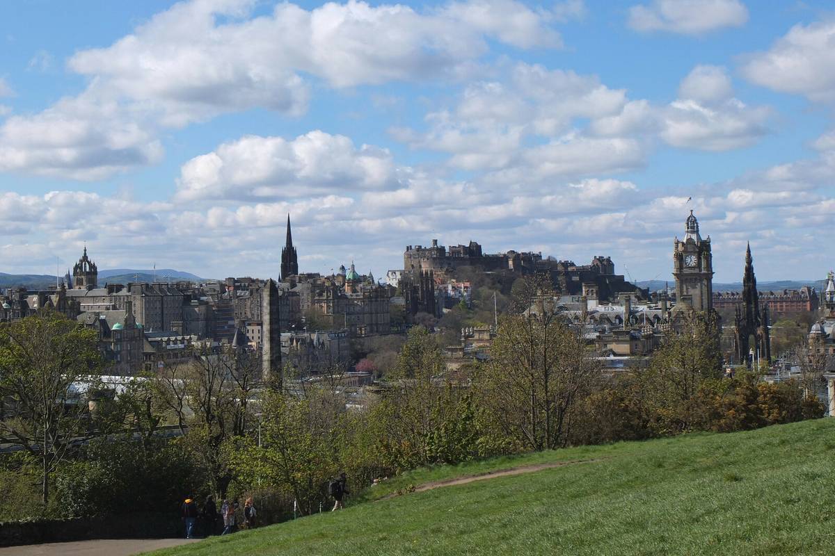 Edinburghs stadssilhuett med kyrktorn, ett klocktorn och borgen på sin klippa, sedd från en grön sluttning på Calton Hill under vita moln