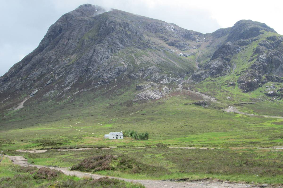 Det spetsiga, klippiga berget Buachaille Etive Mòr reser sig över en grön dalbotten med en grusstig och ett litet vitt hus vid foten av berget