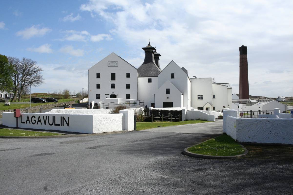 Lagavulins vitkalkade destilleribyggnader med pagodtak och en hög skorsten, med namnet LAGAVULIN målat på en vit mur vid vägen