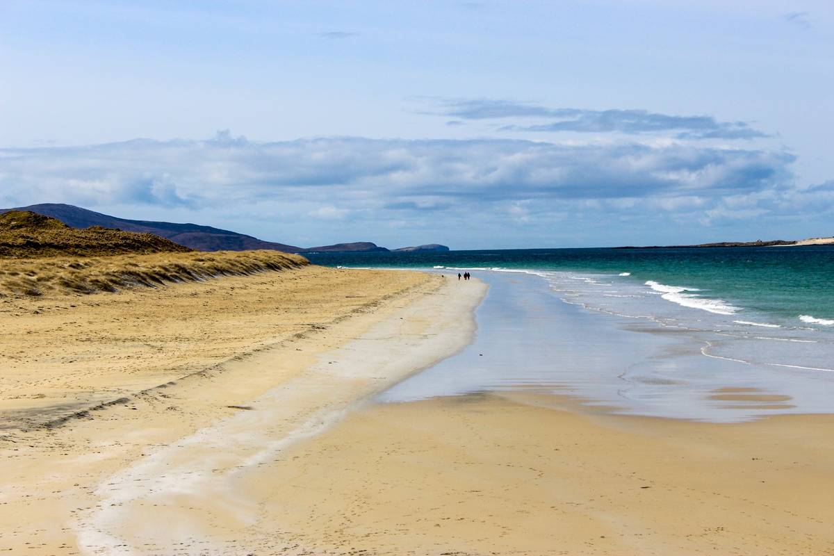 En bred, ljus sandstrand vid Luskentyre på Harris där små vågor sköljer in från ett turkost hav, med en grön kulle och blå berg i fjärran