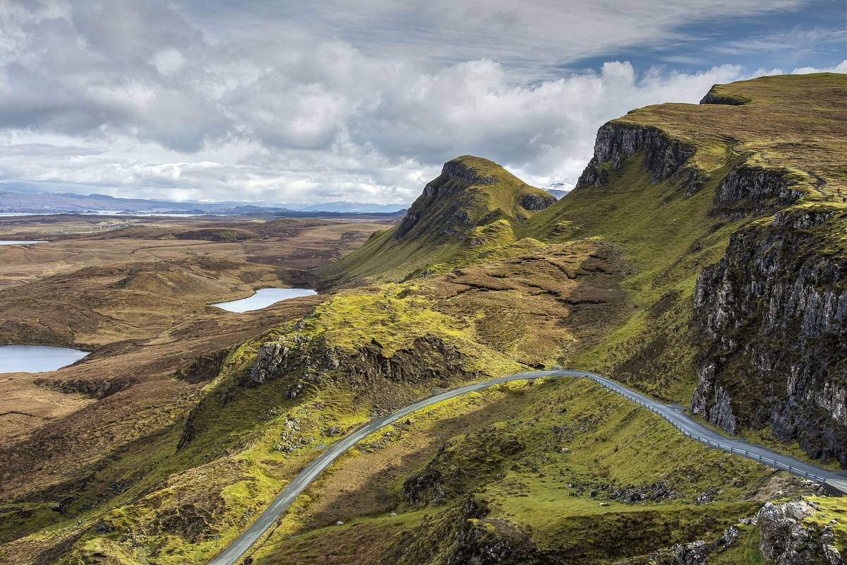 En smal väg slingrar sig genom det gröna, kuperade landskapet vid Quiraing på Isle of Skye, med branta klippor, små sjöar och havet i fjärran