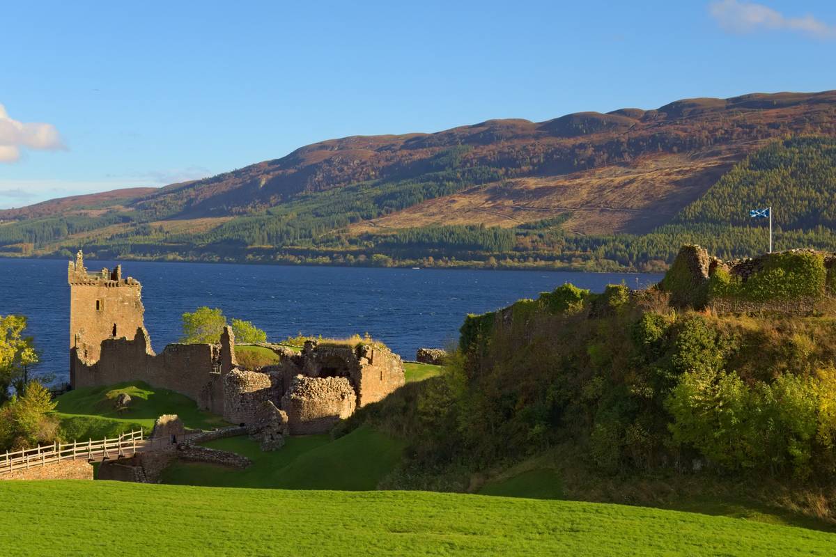 Ruinen Urquhart Castle med ett stentorn på en grön udde vid den blå sjön Loch Ness, med höstfärgade kullar på andra sidan under en klarblå himmel