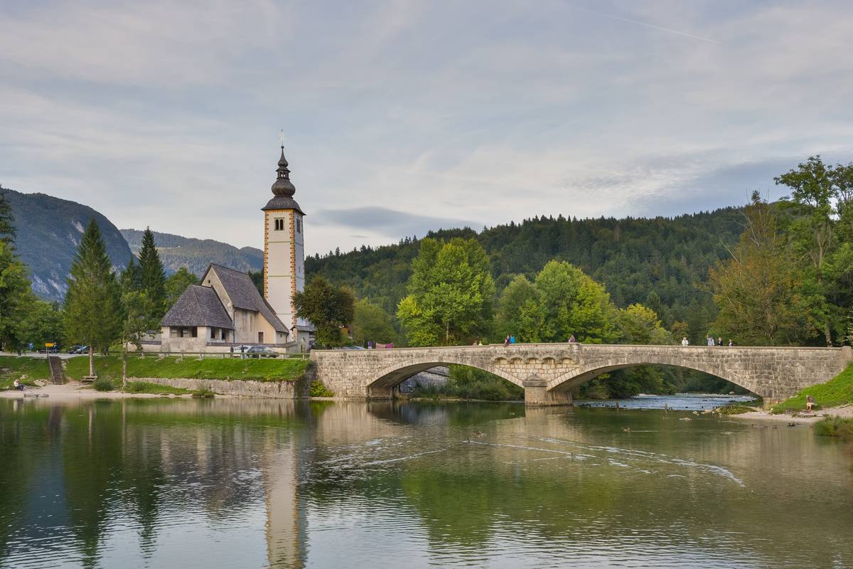 En liten stenkyrka med högt klocktorn och en välvd stenbro speglar sig i det stilla vattnet vid Bohinjsjön, omgiven av gröna skogsklädda berg