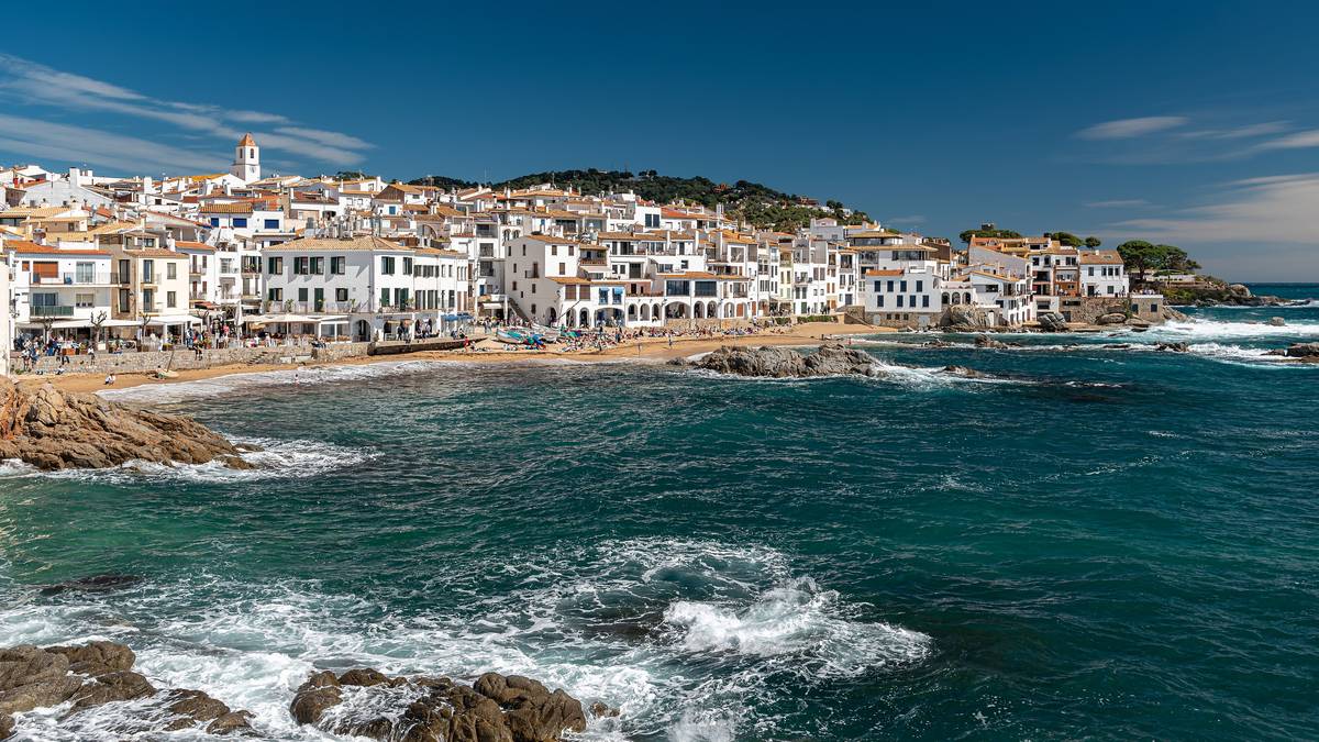 Fiskeläget Calella de Palafrugell på Costa Brava med vita hus längs en liten sandstrand och vågor som bryter mot klipporna i förgrunden
