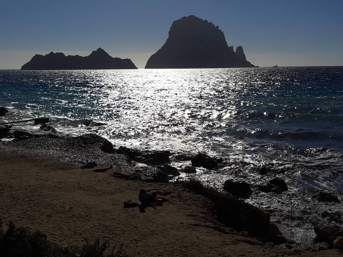 Klippön Es Vedrà i siluett mot eftermiddagssolen, som glittrar på havet utanför den steniga stranden vid Cala d'Hort på Ibiza