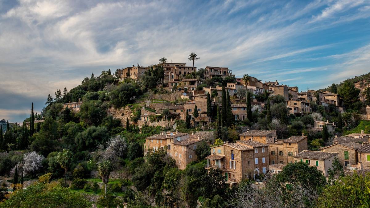 Bergsbyn Deià på Mallorca med honungsfärgade stenhus som klättrar upp för en grön kulle, med cypresser och palmer under en blå himmel