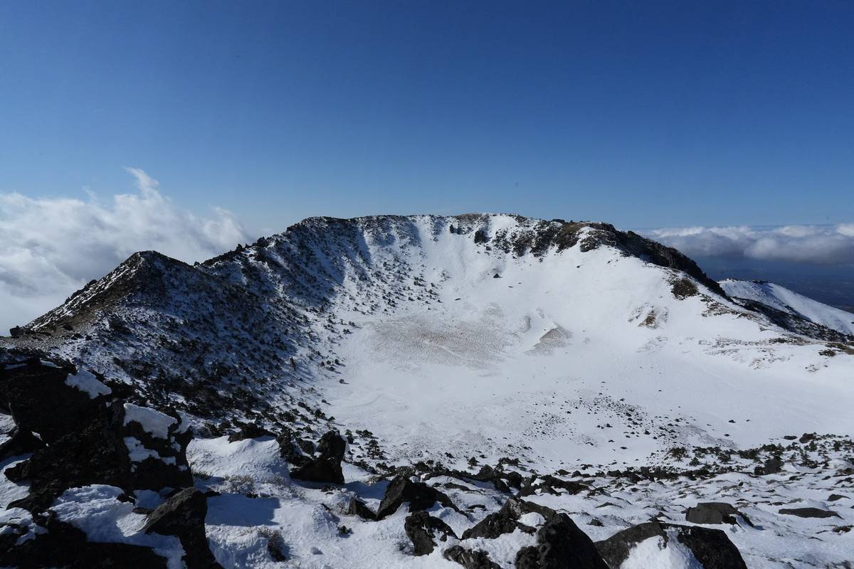 Den snötäckta kratern Baengnokdam på toppen av Hallasan på Jeju, med svarta lavastenar i förgrunden och moln under en blå himmel