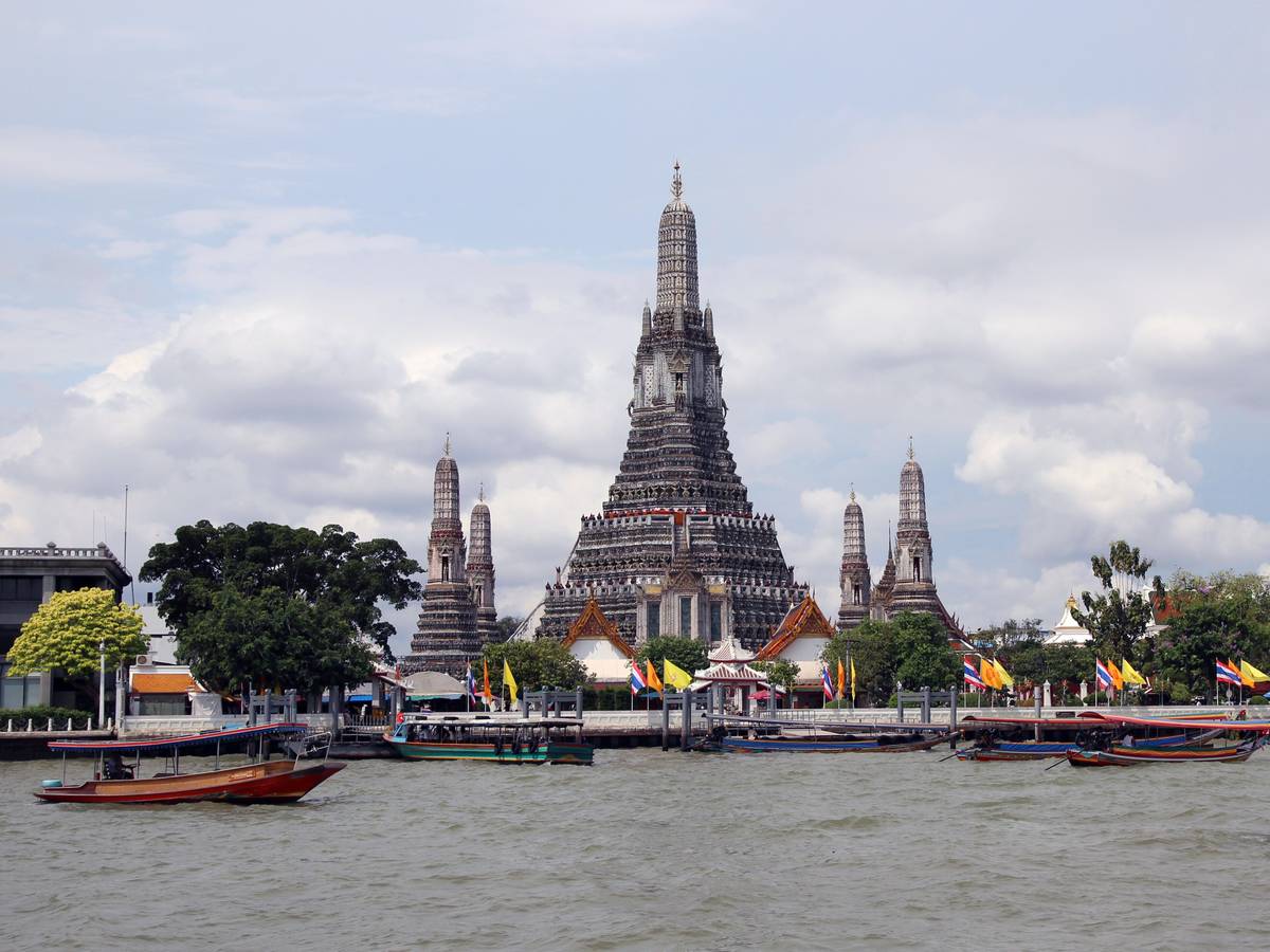 Wat Aruns höga, utsmyckade tempeltorn reser sig vid floden Chao Phraya i Bangkok, med en båt i förgrunden
