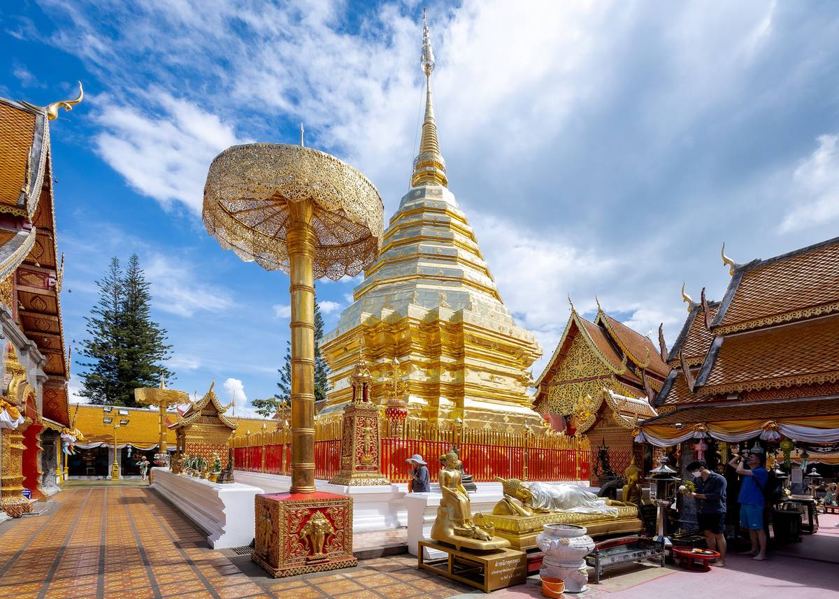 Den gyllene stupan i templet Wat Phra That Doi Suthep med ett guldparasoll framför och tempelbyggnader runt omkring under blå himmel