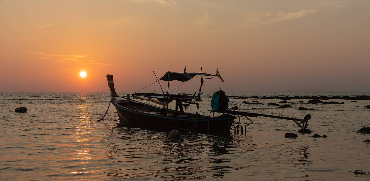 Siluetten av en långsvansbåt i det grunda vattnet vid Klong Khong på Koh Lanta medan solen går ner i en orange himmel