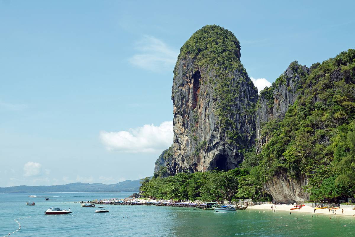 En hög, grönklädd kalkstensklippa reser sig över den ljusa stranden Phra Nang i Railay, med turkost vatten och båtar framför