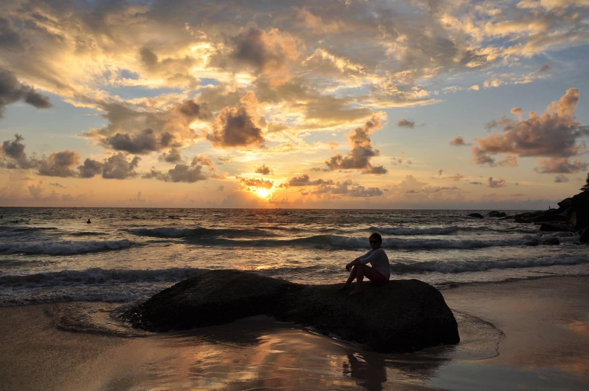 Solnedgång över havet vid Kata Noi på Phuket, med en person som sitter på en klippa i strandkanten under ett orangefärgat molntäcke