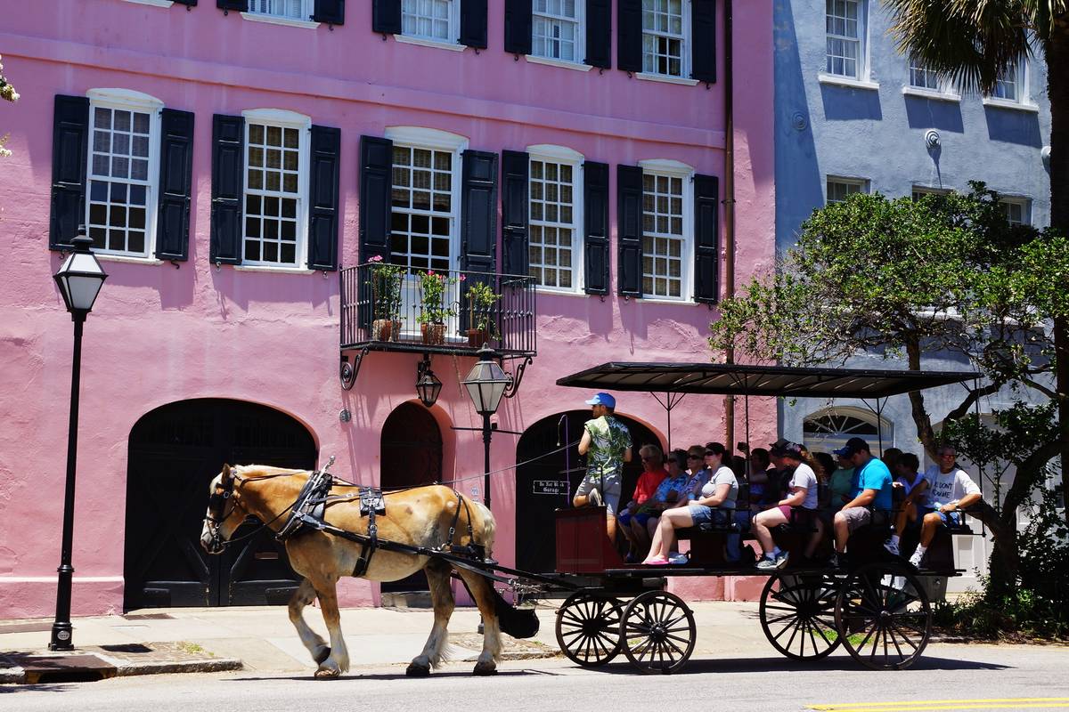 En hästdragen vagn med turister passerar ett rosa och ett blått historiskt hus med mörka fönsterluckor på Rainbow Row i Charleston