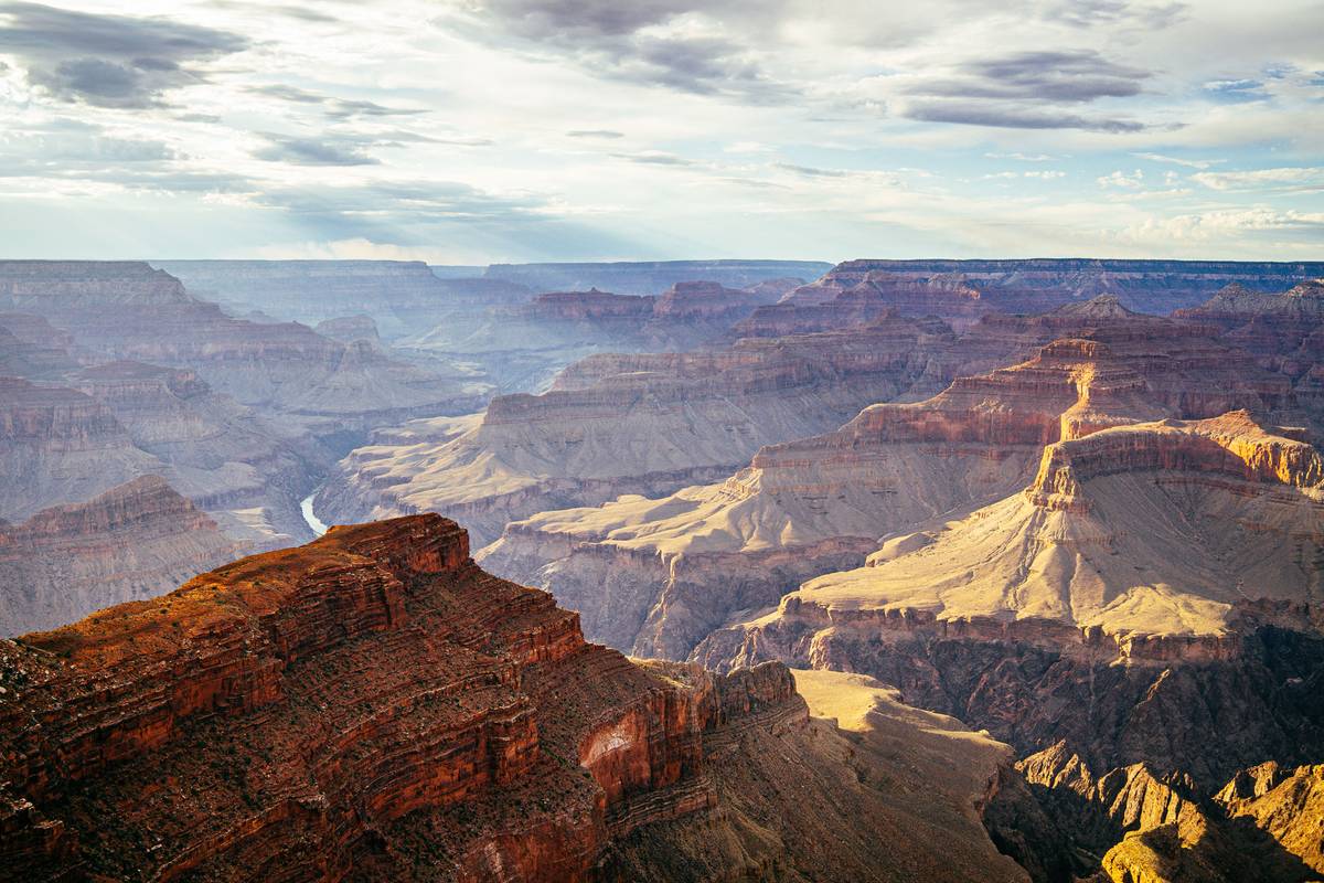 Grand Canyons skiktade klippor i rött, orange och lila i eftermiddagsljus, med Coloradofloden djupt nere i kanjonen och moln på himlen