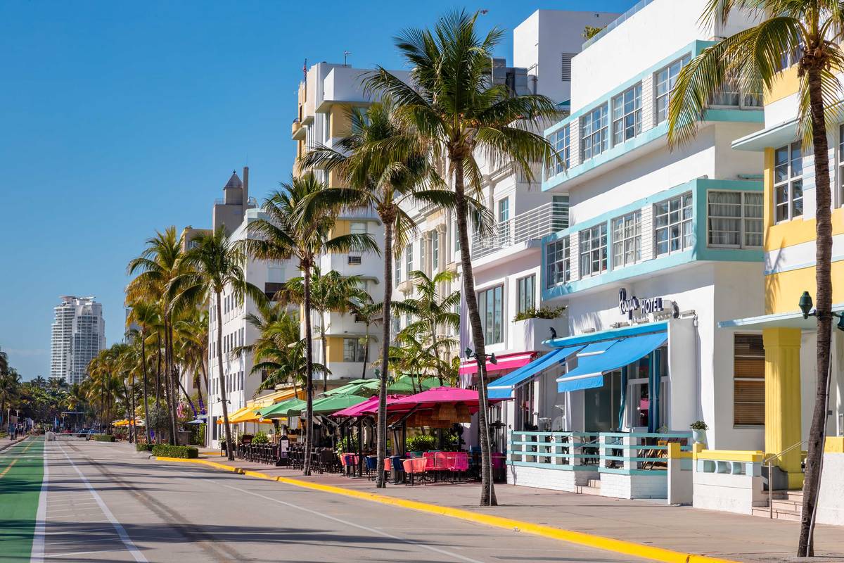 Pastellfärgade art déco-hotell med markiser och uteserveringar längs Ocean Drive i Miami Beach, kantade av höga palmer under en blå himmel