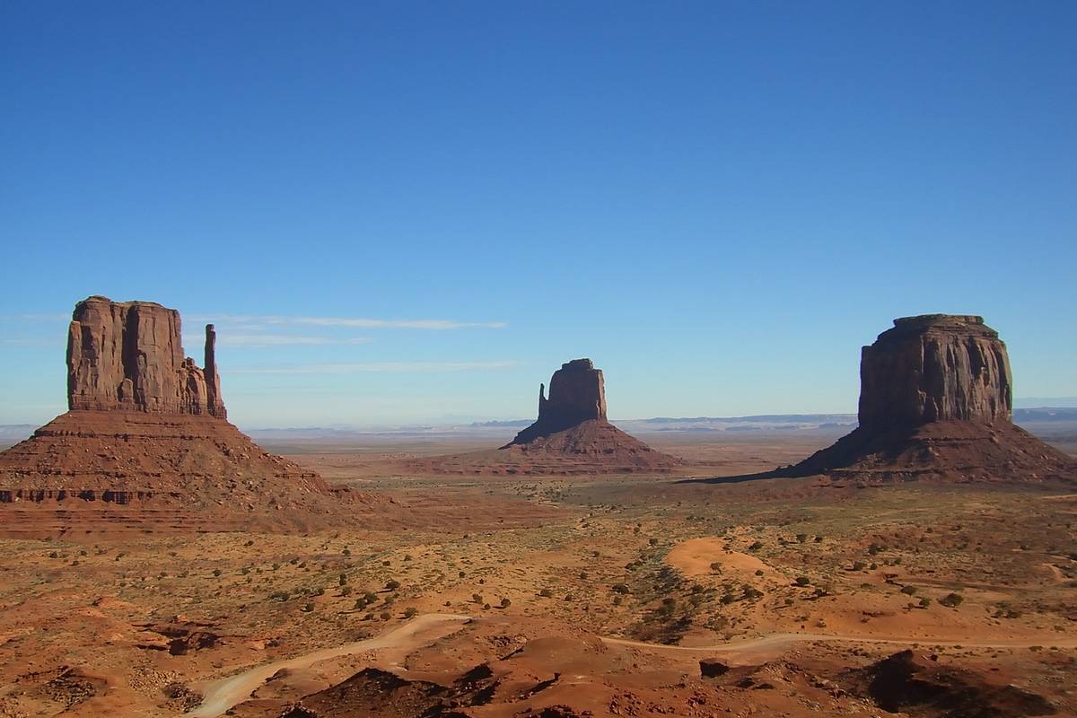 De rödbruna klippformationerna i Monument Valley reser sig ur en platt öken med röd sand och låga buskar under en djupblå himmel