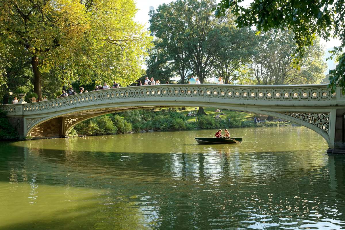 Den ljusa gjutjärnsbron Bow Bridge välver sig över den gröna sjön i Central Park, med en roddbåt på vattnet och lummiga träd runt omkring