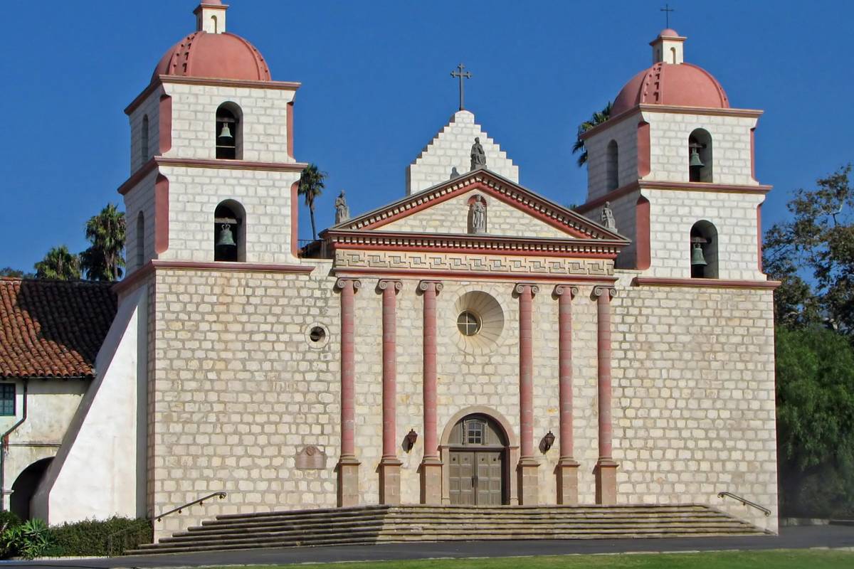 Den ljusa stenfasaden på Old Mission Santa Barbara med två klocktorn med rosa kupoler och pelare framför, under en klarblå himmel