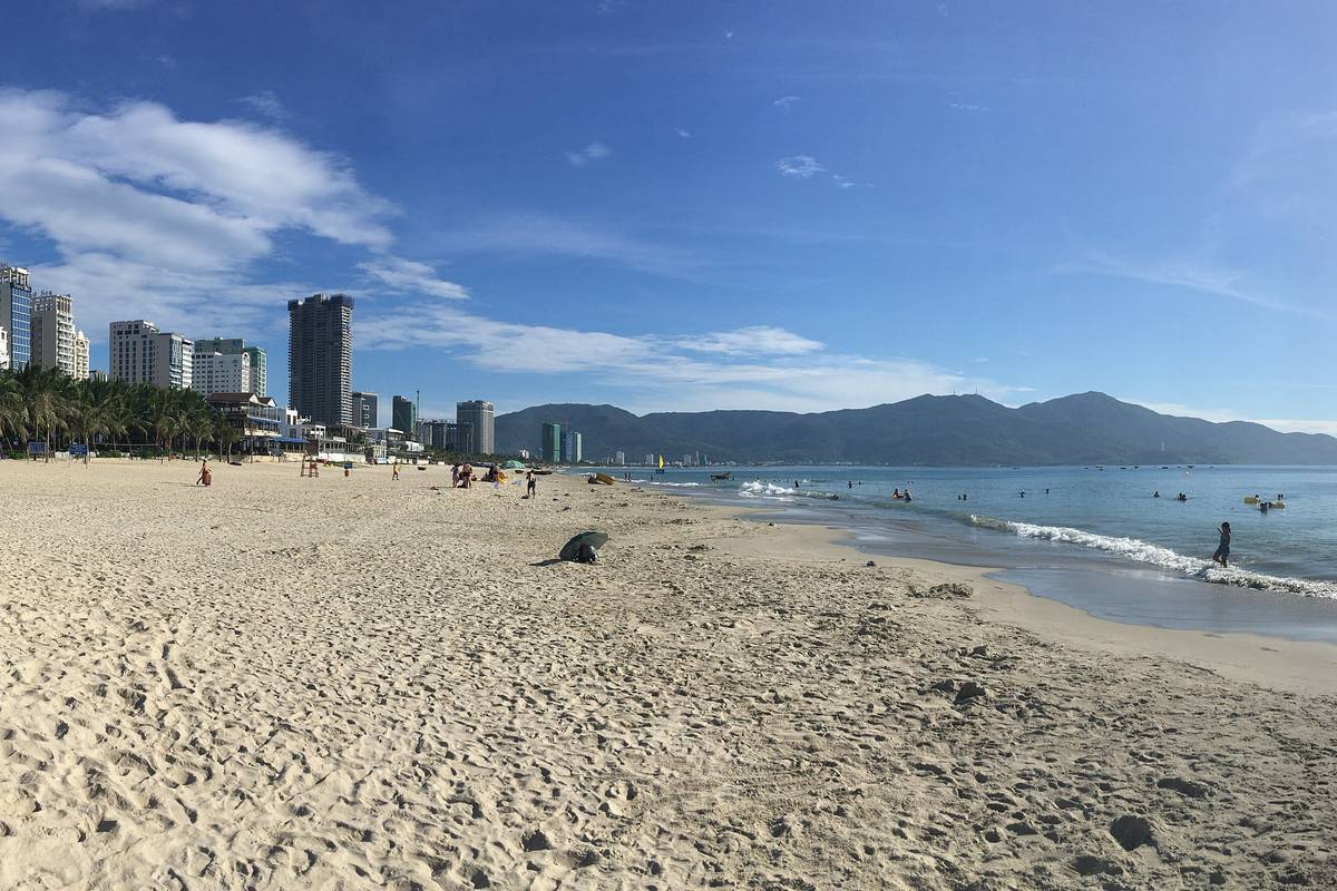 Bred ljus sandstrand vid My Khe i Da Nang med vågor som rullar in, höghus vid strandkanten och den gröna halvön Son Tra i bakgrunden