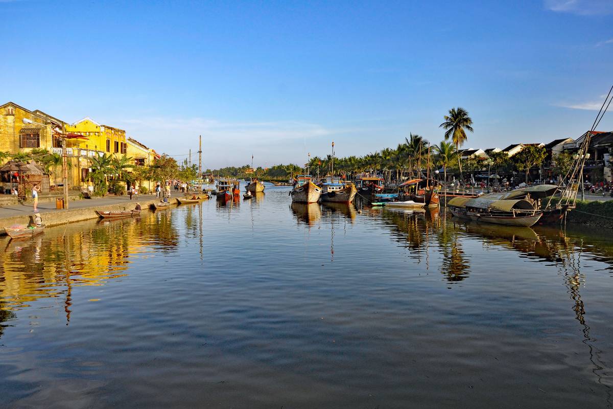 Gula hus och palmer längs Thu Bon-floden i Hoi An, med båtar vid kajen som speglar sig i det stilla vattnet under klarblå himmel