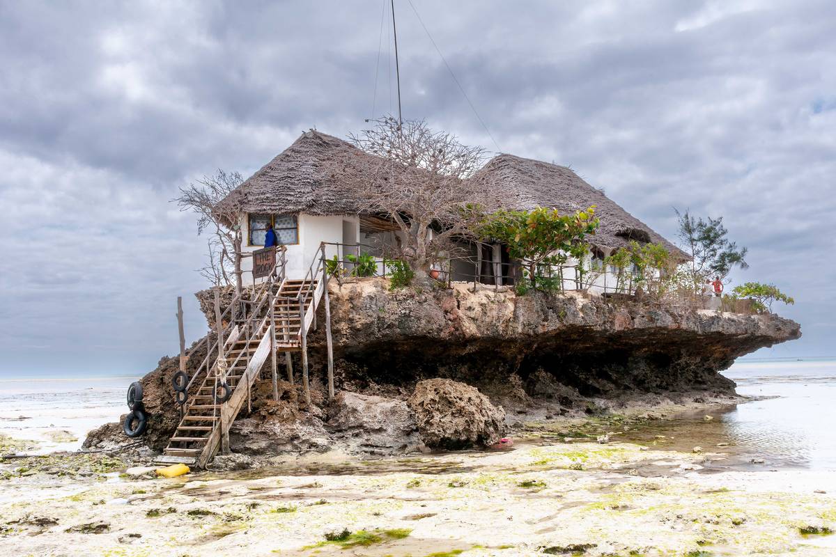 Restaurangen The Rock med halmtak och trätrappa på en klippa i det grunda vattnet utanför stranden vid lågvatten